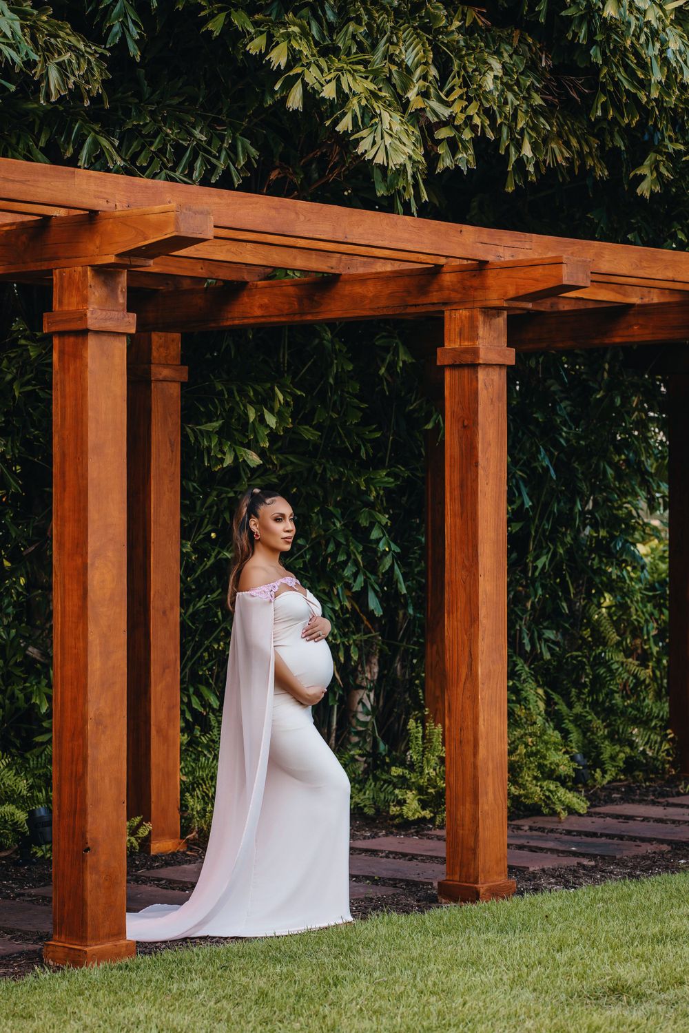 Silhouette of pregnant woman in white dress under garden pergola.