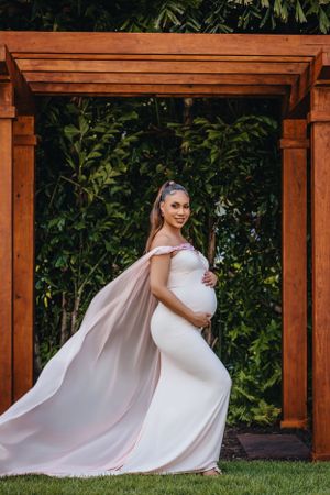 Dramatic maternity portrait with flowing cape under wooden pergola.