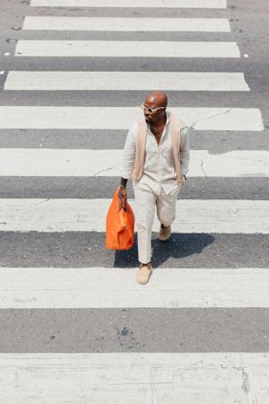 Bird's eye view of pedestrian in neutral tones crossing striped crosswalk with orange shopping bag.