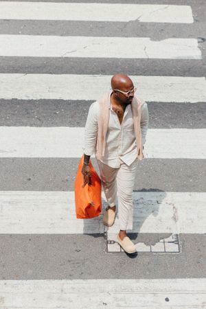 Overhead shot of person in light-colored outfit crossing street with bright orange shopping bag.