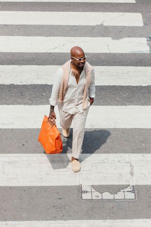 Stylishly dressed person crossing zebra crossing while carrying orange tote bag in urban setting.