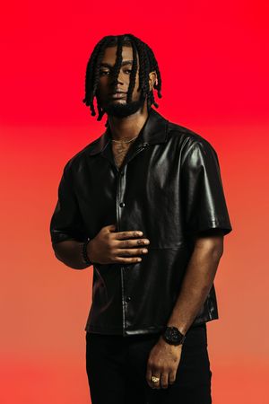 Stylish figure in black leather attire poses against vibrant red gradient background wearing sunglasses.