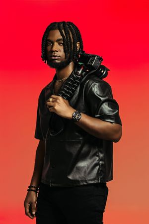 Stylish photographer in leather vest poses with professional camera against vibrant red background.
