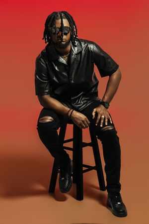 Artistic studio shot of someone seated on stool wearing all black against red-orange background.