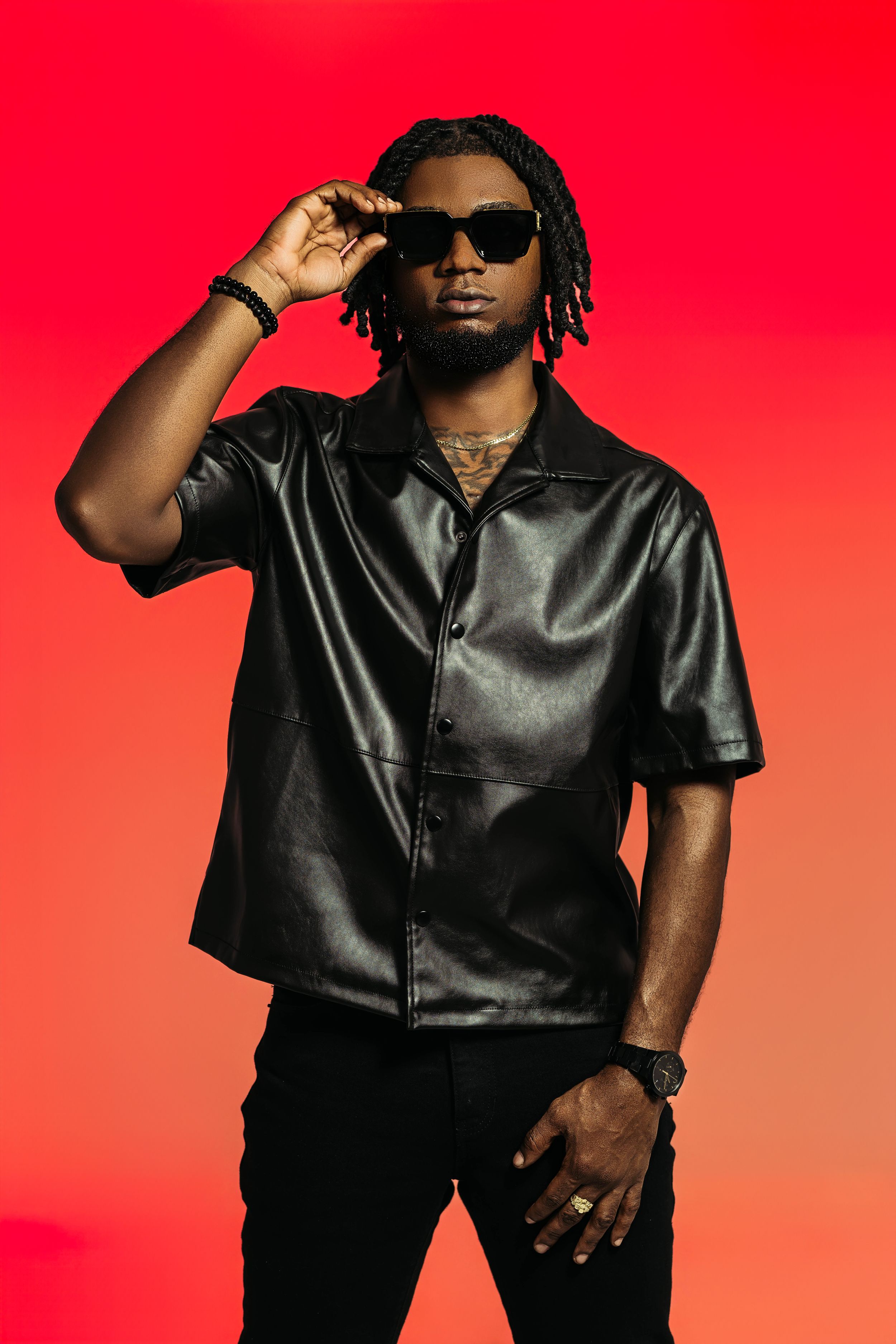 Urban fashion portrait with person in dark leather outfit and sunglasses against red backdrop.