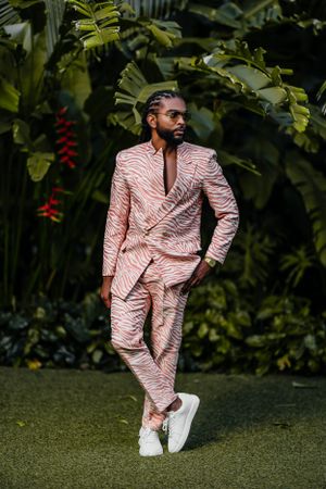 Stylish model in pink textured suit and white sneakers poses against tropical foliage.