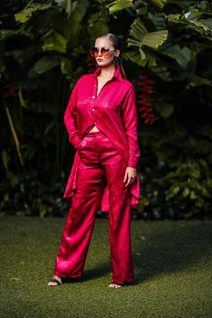 Model in bright red coordinated outfit poses with sunglasses against green foliage.