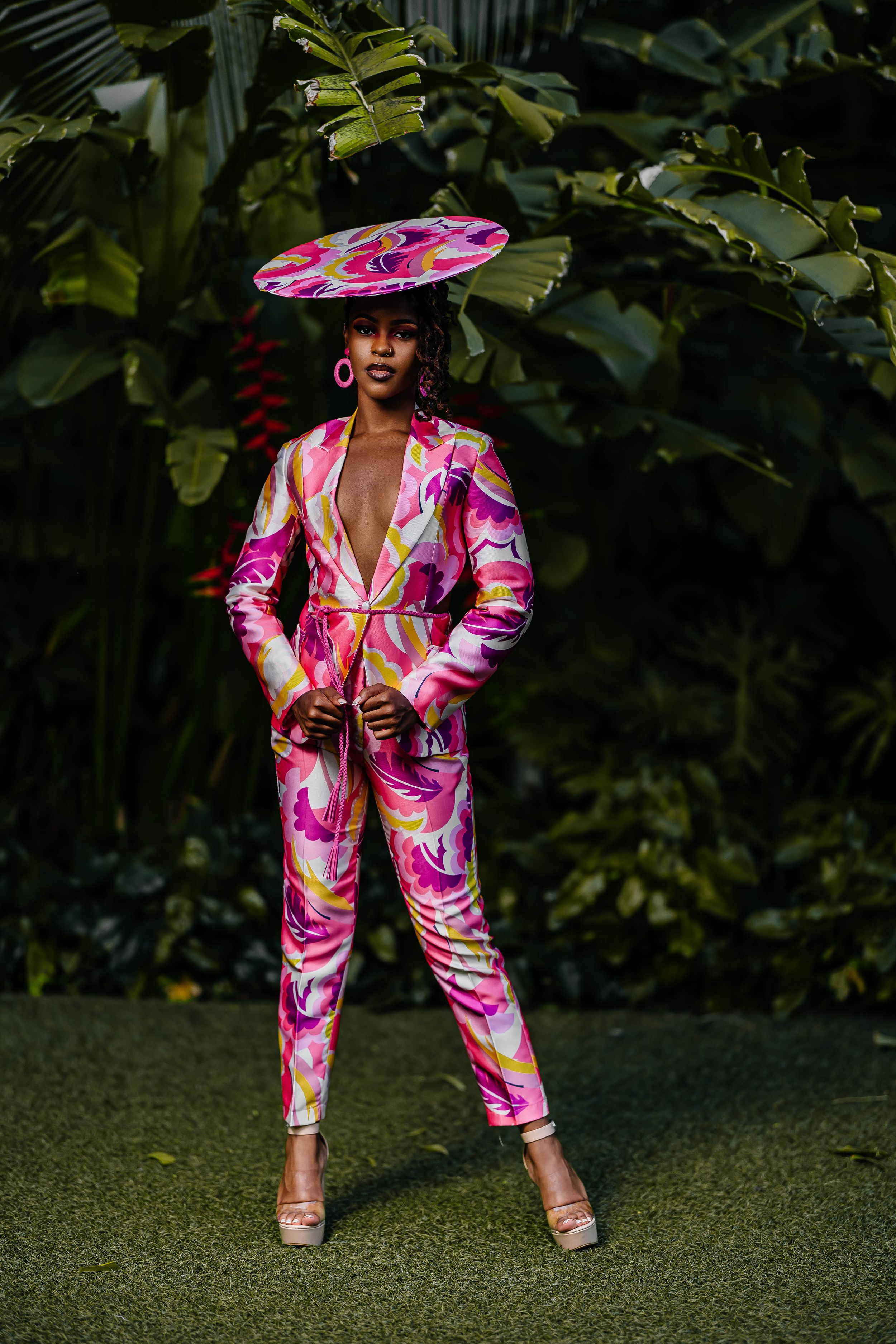 Model in pink floral jumpsuit and statement hat poses dramatically in garden setting.