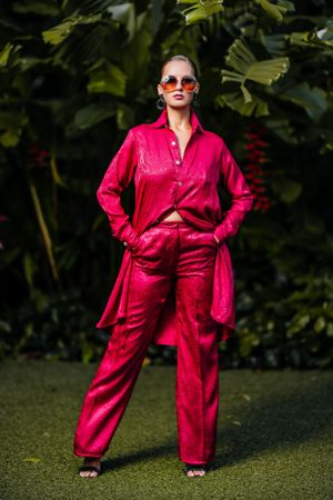Fashion model in vibrant red silk pantsuit and sunglasses poses against tropical foliage backdrop.