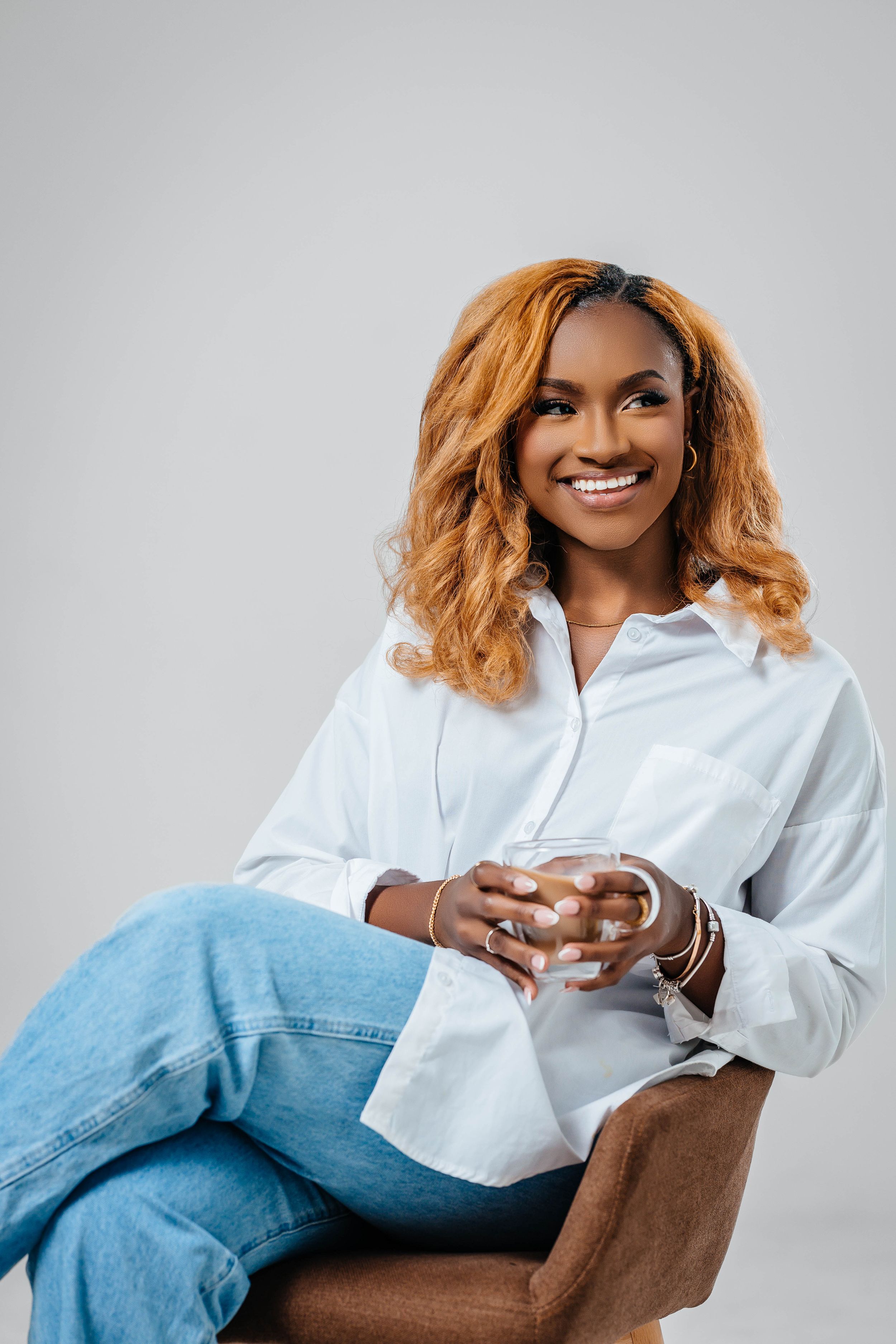 A relaxed portrait showing someone in a white shirt and jeans seated comfortably against a gray background.