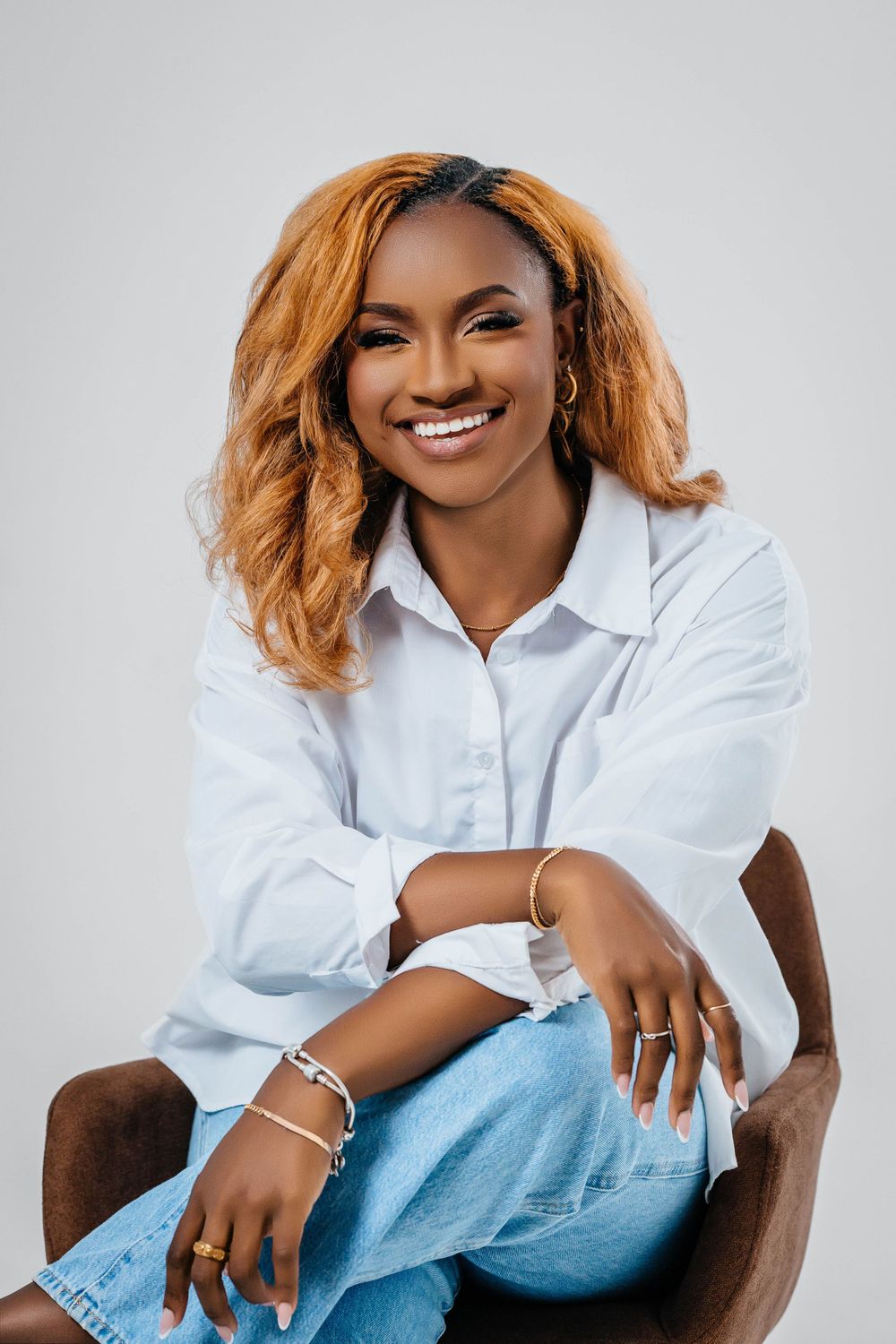 A professional portrait of someone in a white button-down shirt and jeans sitting casually in a brown chair.