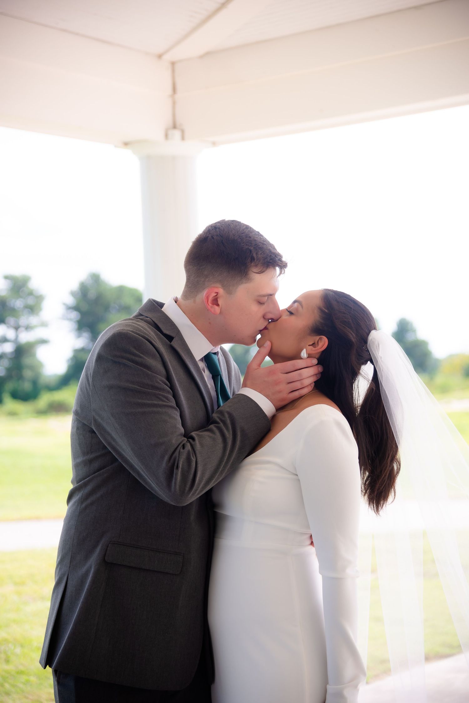 A couple shares a romantic kiss during their wedding ceremony under a covered outdoor venue with green scenery.
