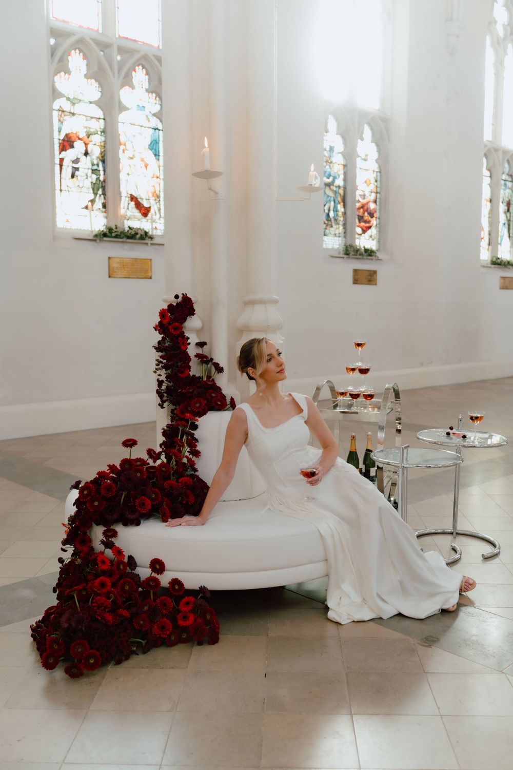 A bride in a flowing white gown sits elegantly beside deep red floral arrangements in a light-filled chapel setting.