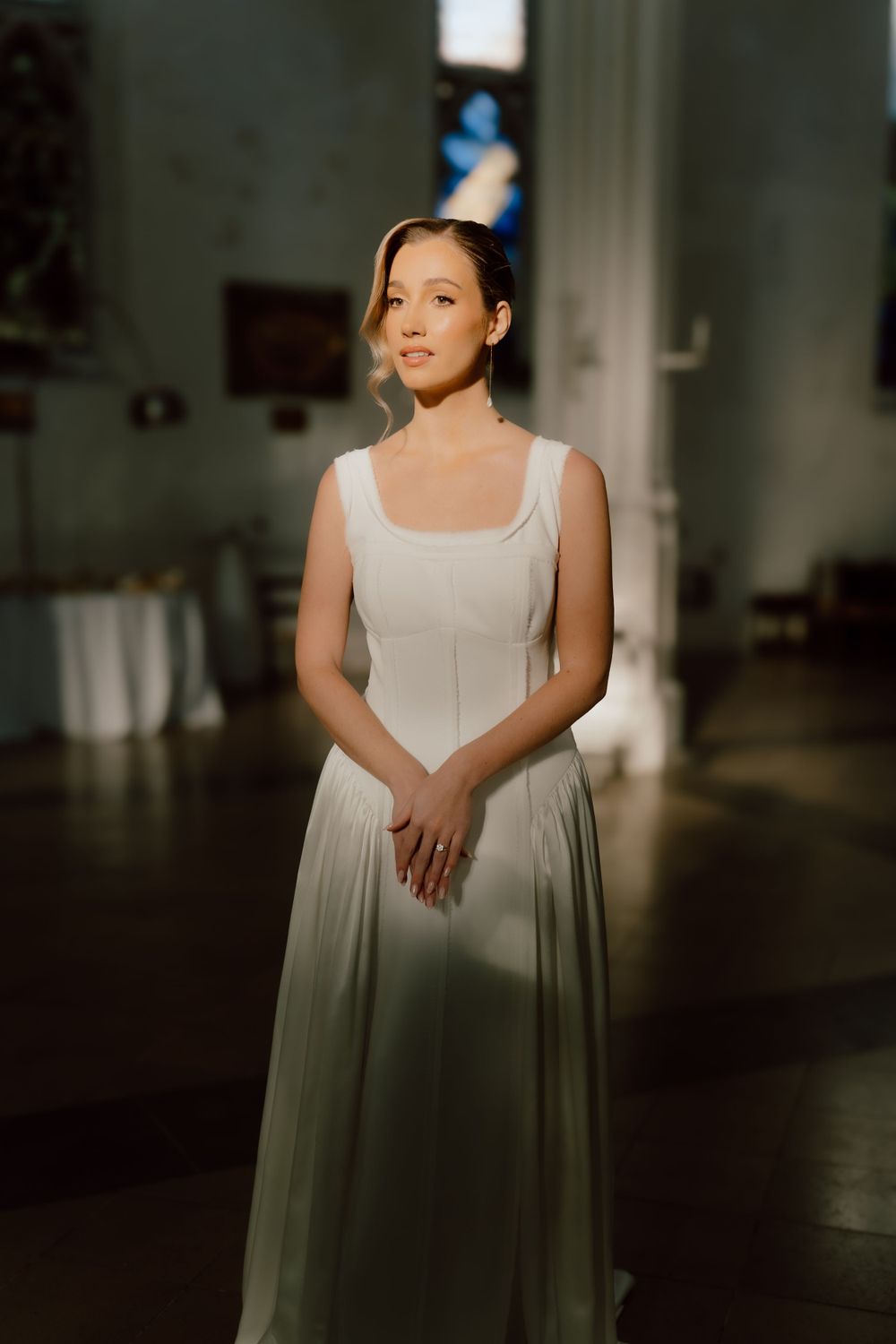 Figure in a flowing floor-length white dress poses elegantly in soft dramatic lighting indoors.