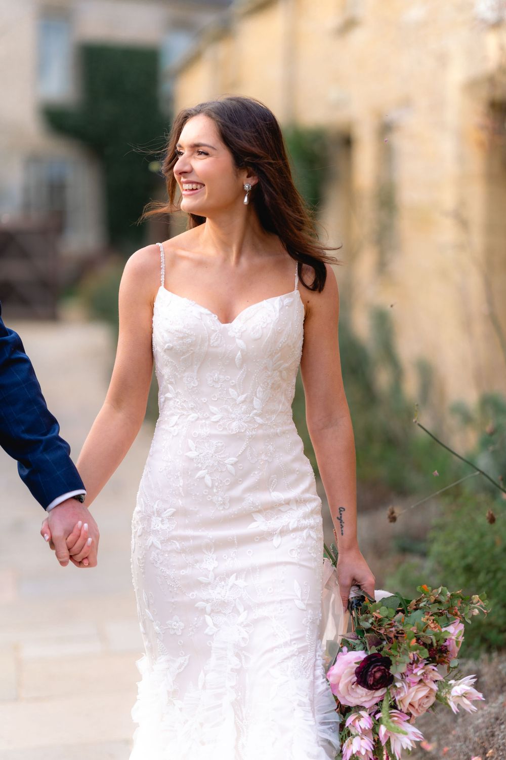 Bride in fitted lace dress holding bouquet while walking along Mediterranean villa pathway.