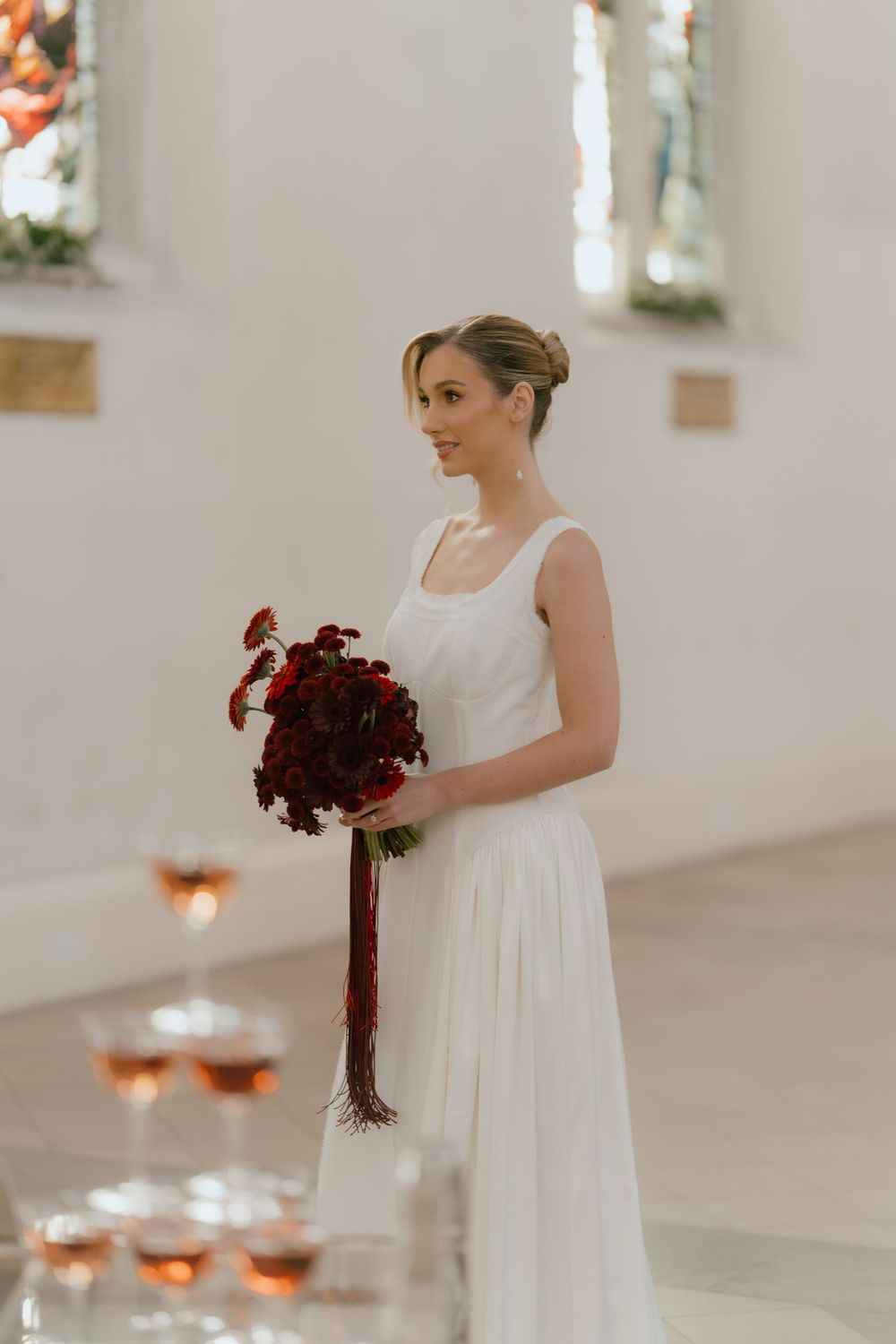 Bride in simple white dress holds deep red bouquet in elegant church setting.