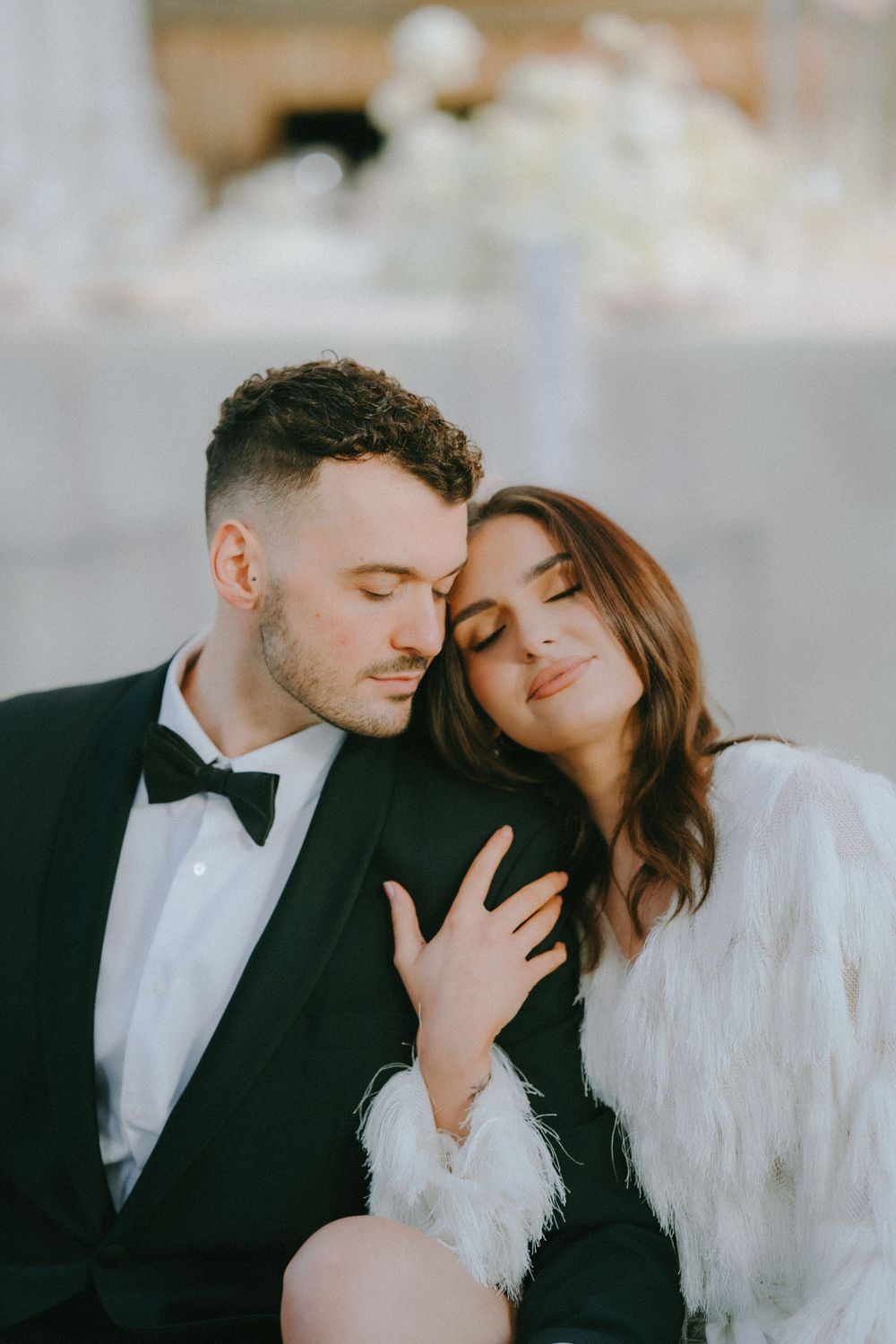 Romantic wedding couple embraces in formal attire with feathered details against soft background.