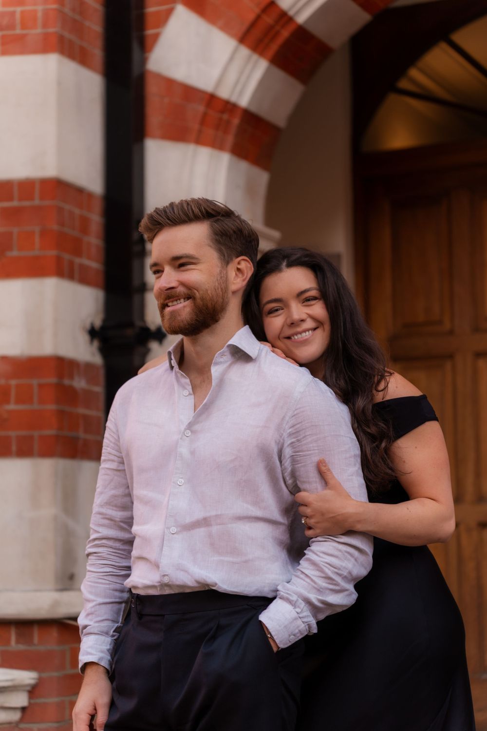 Couple sharing intimate moment outside historic red brick building with striped arches.