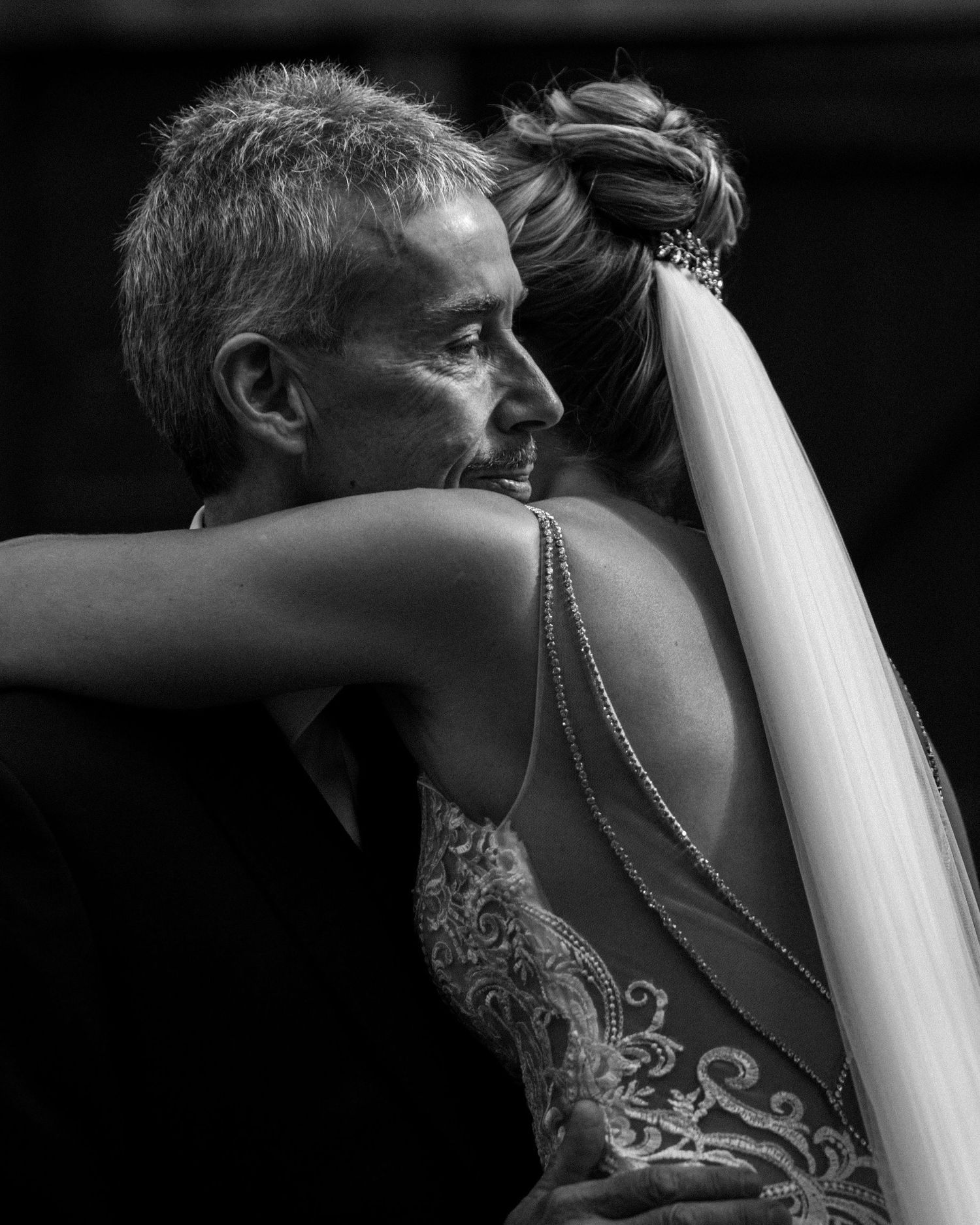 A dramatic black and white portrait showing a father hugging a bride in an embellished dress with delicate back details in Nova Scotia