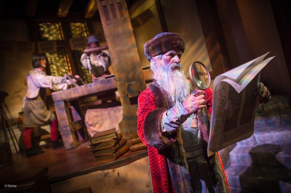 A costumed performer in a red robe reads from a large book in a dimly lit medieval-themed attraction.
