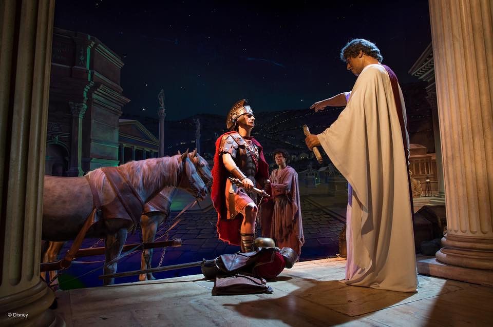 A theatrical stage scene shows a draped figure standing over a kneeling figure, with a decorated horse prop in the background.