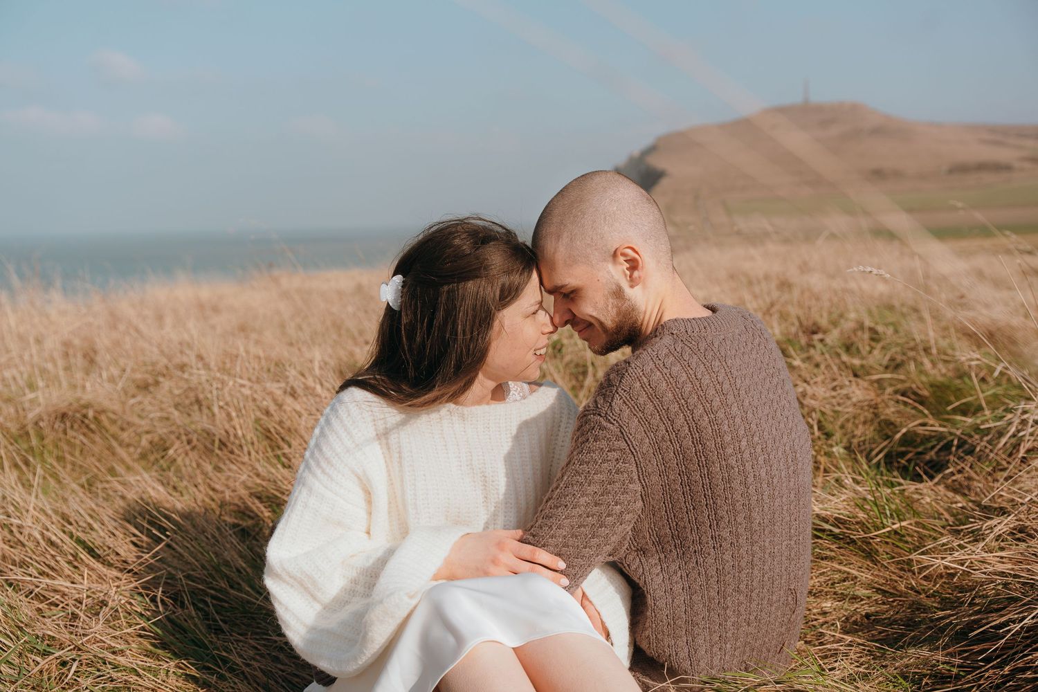 A couple sitting in a golden field of tall grass embrace against a mountain backdrop on a sunny afternoon.