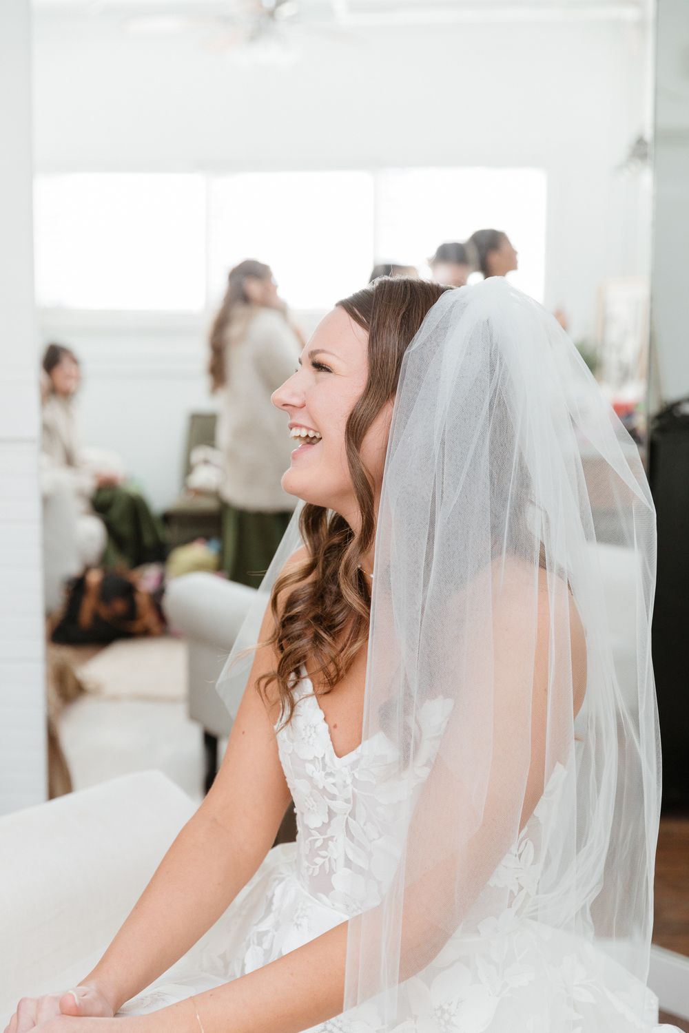A bride in a white wedding dress and veil laughs during preparations.