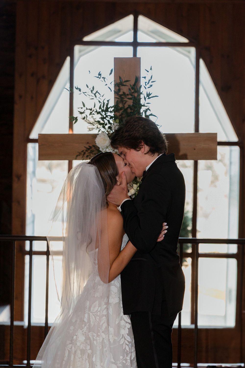 Newlyweds share their first kiss as a married couple in front of a sunlit window.
