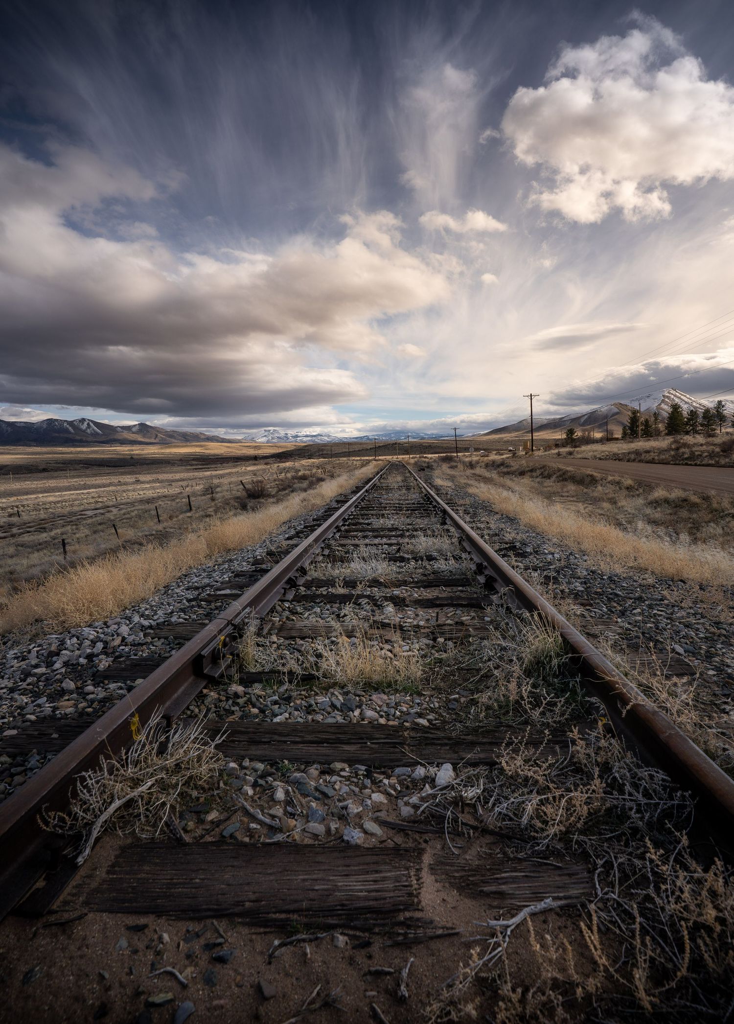 Old railroad tracks lead toward distant mountains under dramatic cloudy skies in a rural desert landscape.