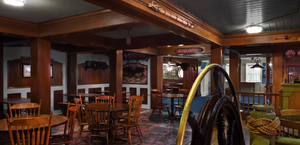 Interior view of an empty restaurant dining room with wooden tables and chairs under warm lighting and exposed ceiling beams.
