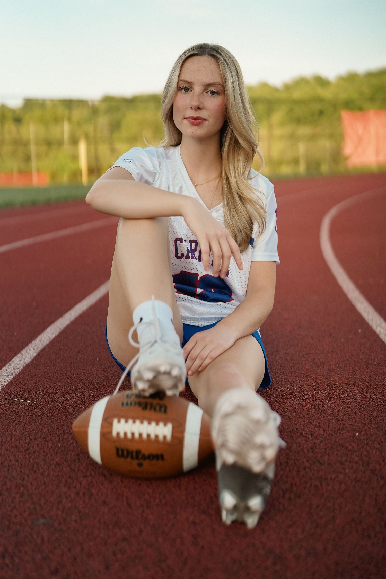 Person sitting on red running track with football and small pet creating playful composition.
