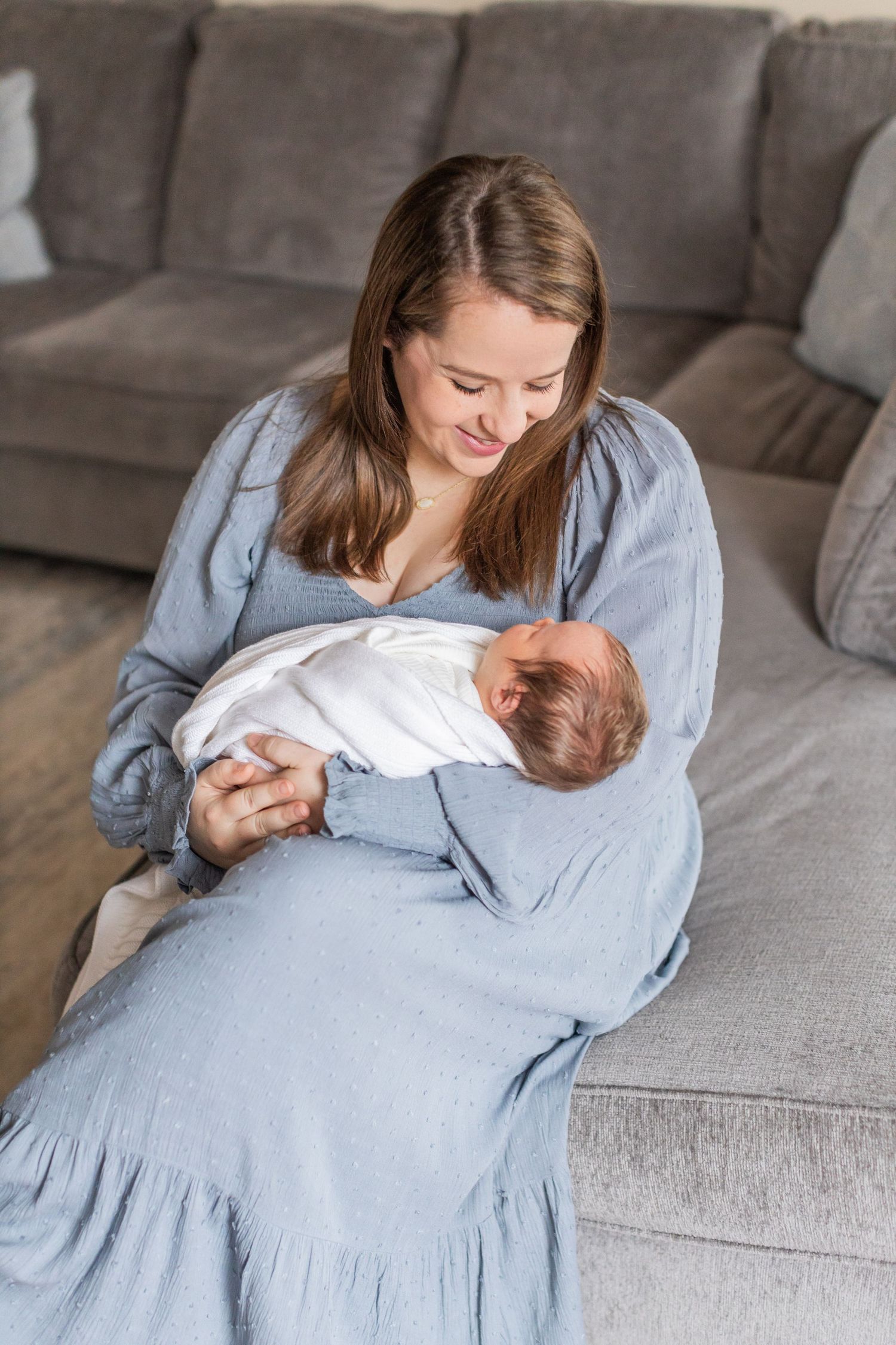 A mother wearing a blue dress cradles and gazes lovingly at her newborn baby wrapped in white on a gray couch.