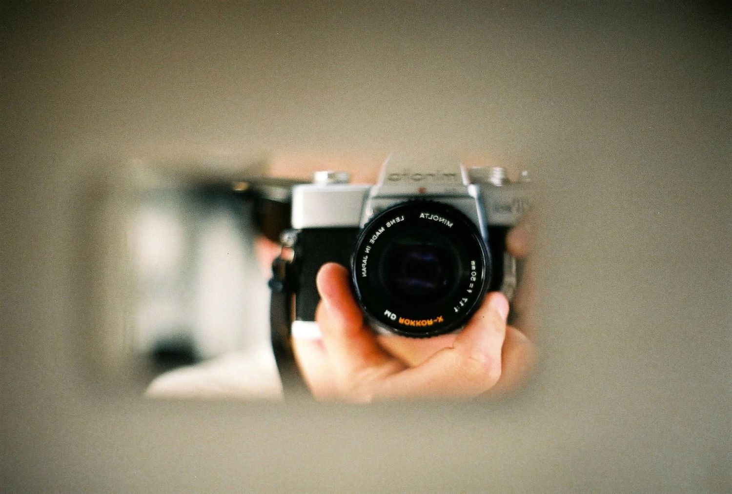 A vintage camera lens being held in front and reflecting in a mirror, creating a recursive self-portrait effect.