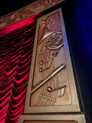 Ornate bronze Art Deco wall relief panels with geometric patterns illuminated by dramatic red stage curtain lighting.