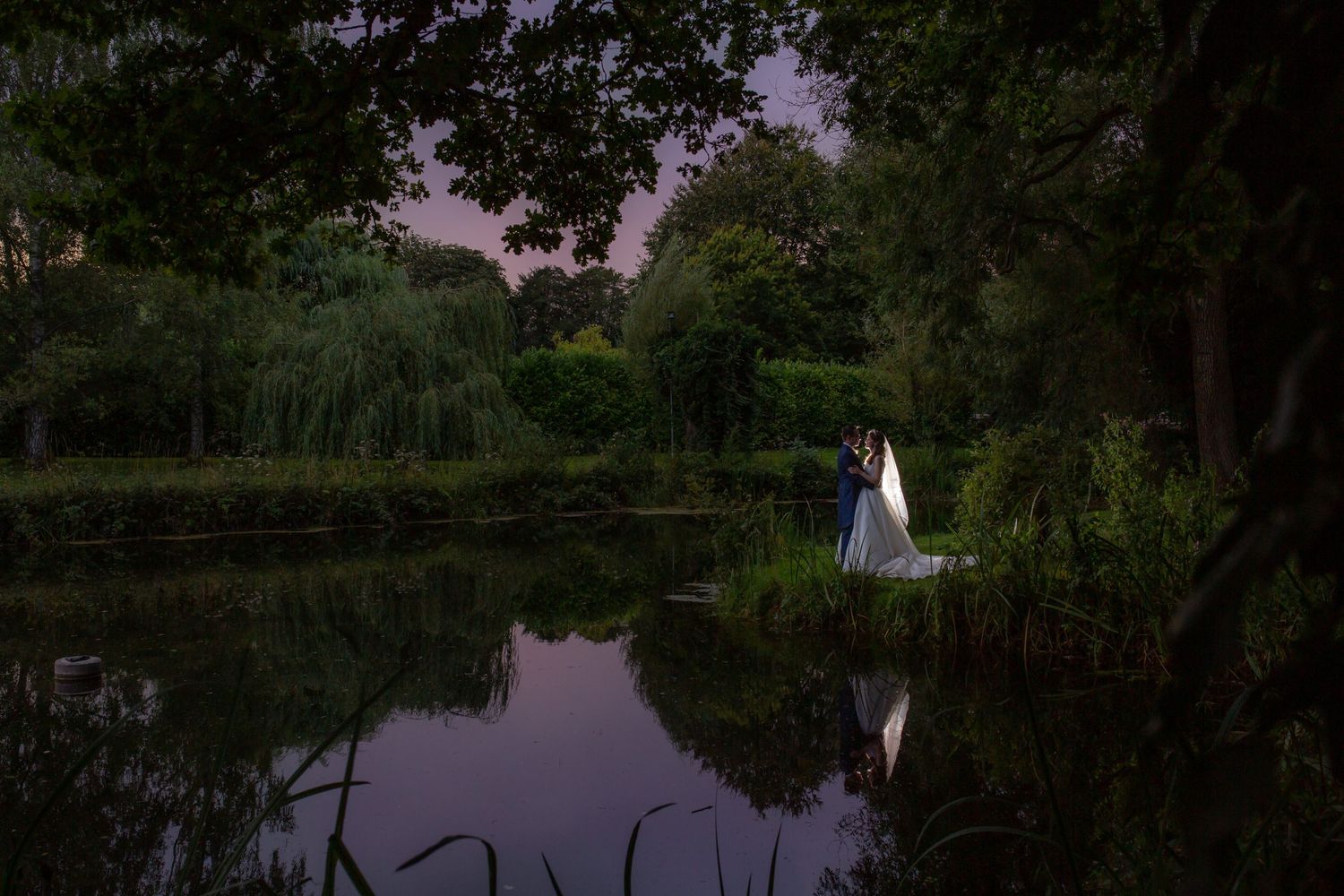 A couple stands near the edge of a tranquil pond at dusk, surrounded by lush trees and greenery for Nova Scotia wedding photographer.