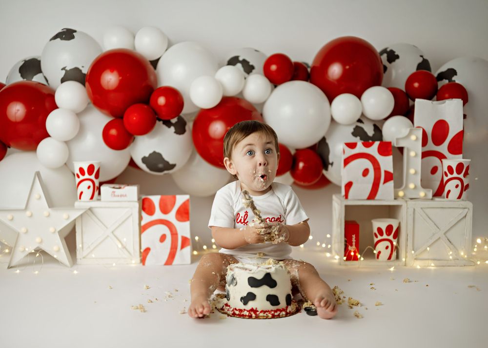 Festive first birthday setup with red and white balloon arch, wrapped presents, and cow print smash cake against neutral backdrop.