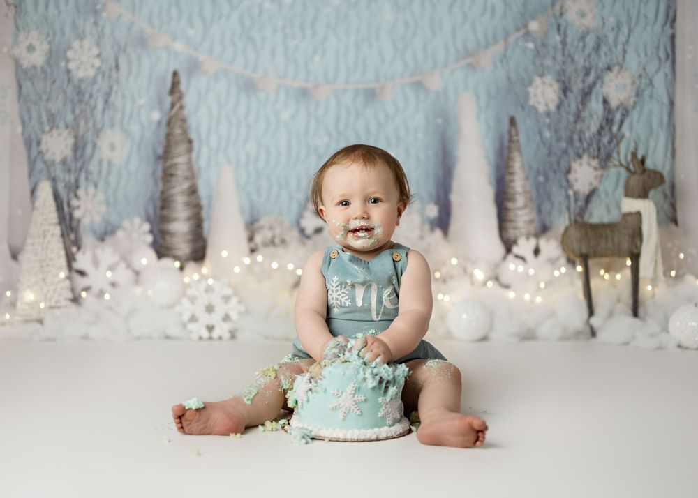 Winter wonderland cake smash photo session with baby in blue outfit sitting with birthday cake on white floor.