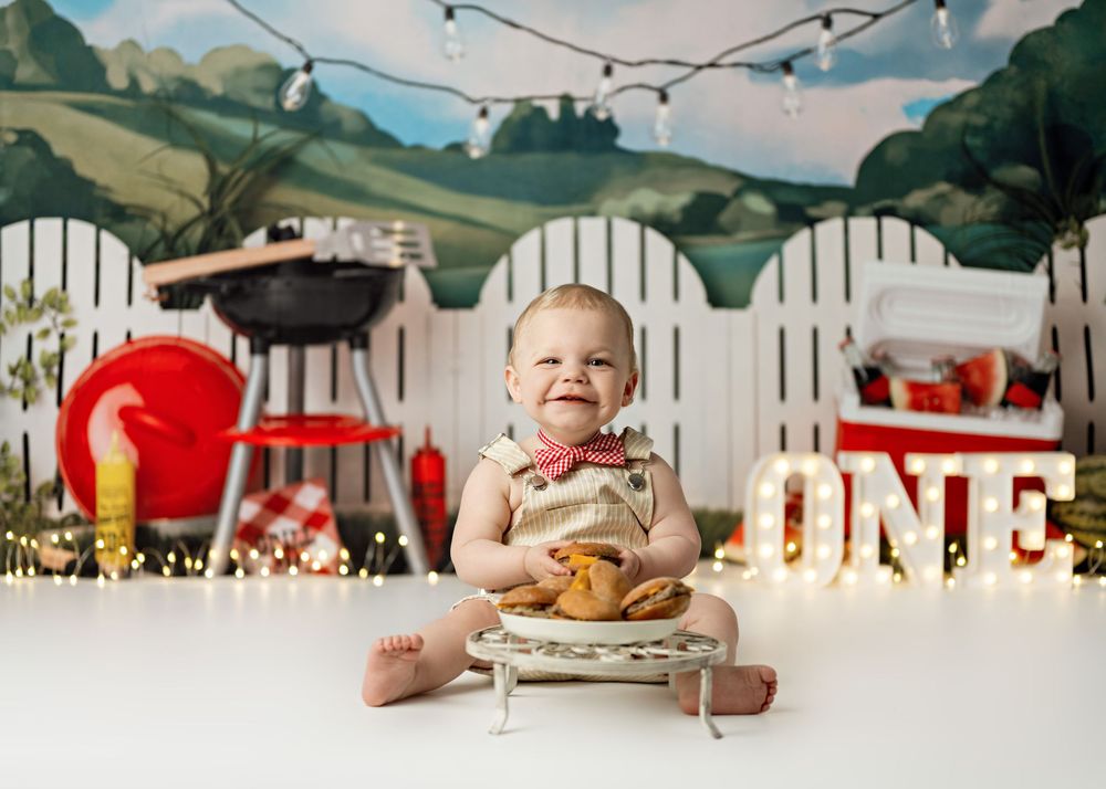 Smiling baby at first birthday cake smash with BBQ backdrop, ONE sign, string lights, and red decorations.