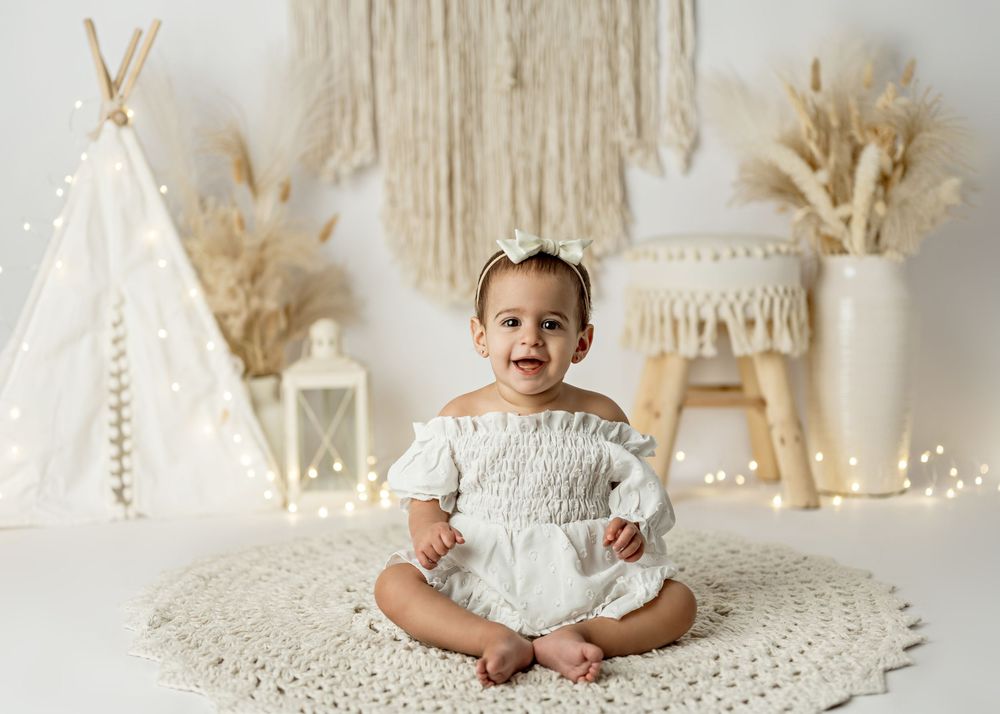 Smiling baby in white romper sits on crochet rug with boho teepee, macrame, and fairy lights backdrop.