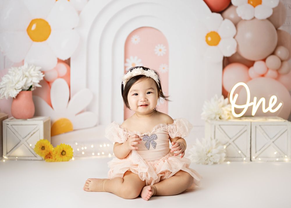 A festive first birthday photo setup with white arches, pink and orange balloons, floral decorations, and the word 'One' displayed.