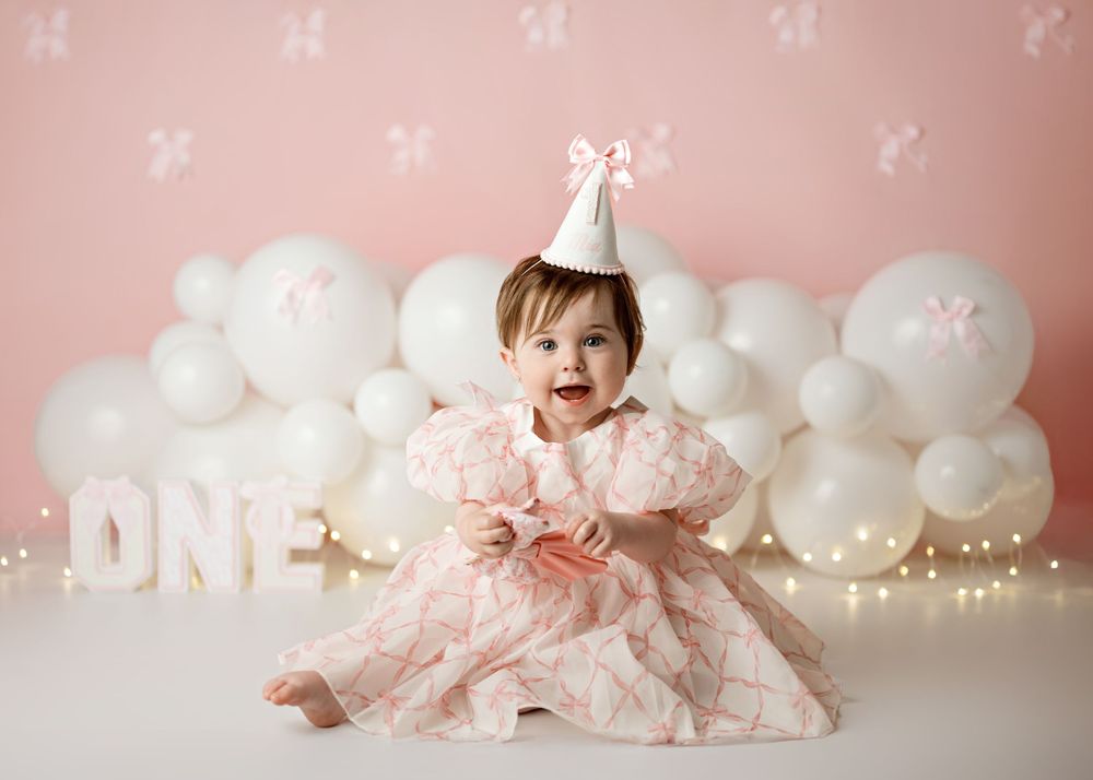A baby in a pink tutu dress celebrates their first birthday with white balloons and party decorations in a studio setting.