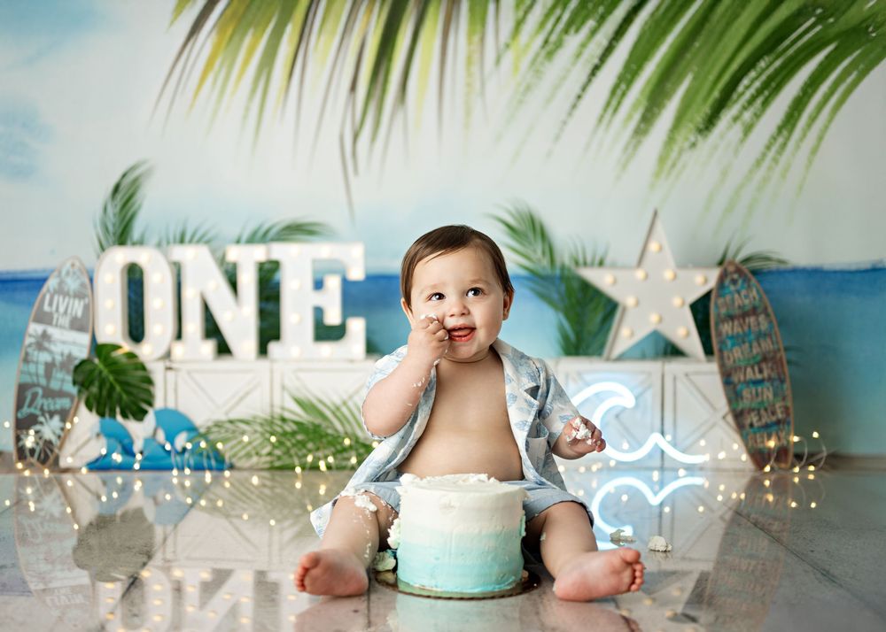 Baby milestone photoshoot with tropical backdrop, palm leaves, and turquoise cake on beach-themed setting.