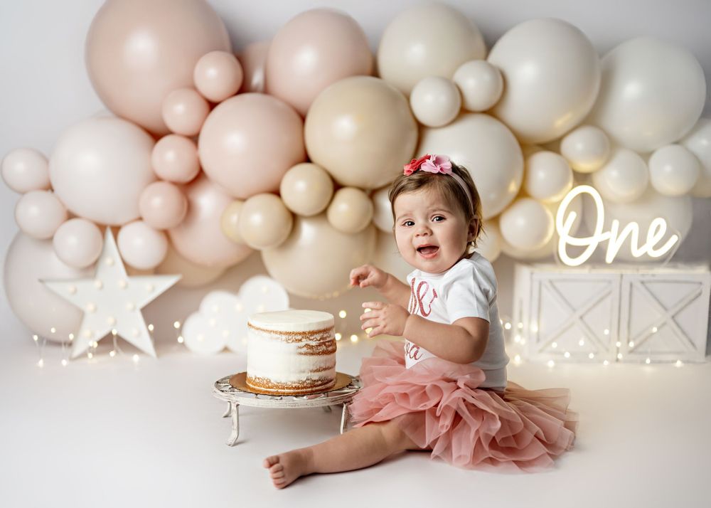A baby in a pink tutu sits on the floor with neutral-toned balloon decorations and a birthday cake for a first birthday photoshoot.