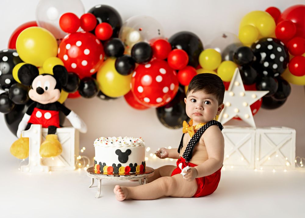 Baby in Mickey Mouse outfit celebrates first birthday with cake, balloons, and Mickey plush in a festive photo shoot.
