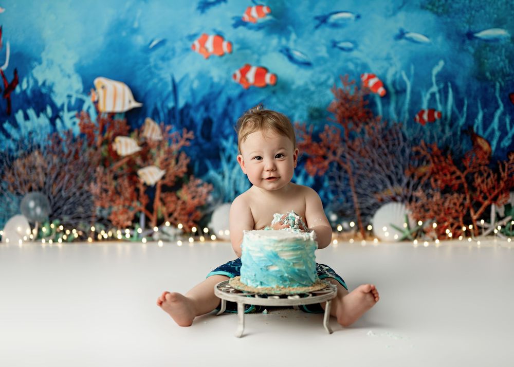 Baby's first birthday cake smash with ocean theme backdrop featuring colorful fish and coral reef decorations.