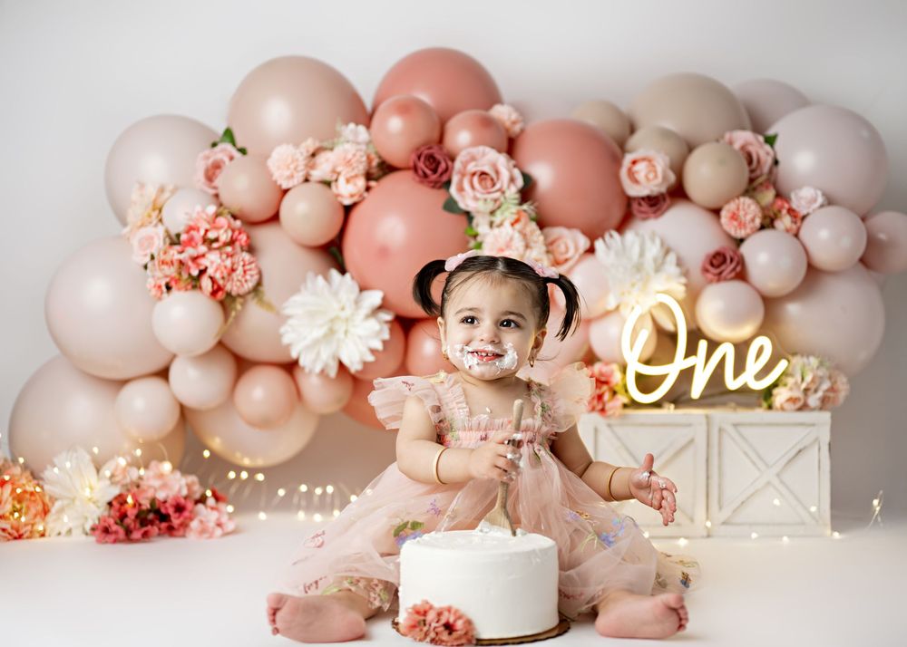 Elegant first birthday setup with blush pink balloon arch, white floral decorations and illuminated 'One' neon sign.