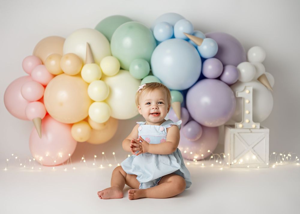 Rainbow pastel balloon arch with baby seated beneath on white backdrop in celebratory setting.