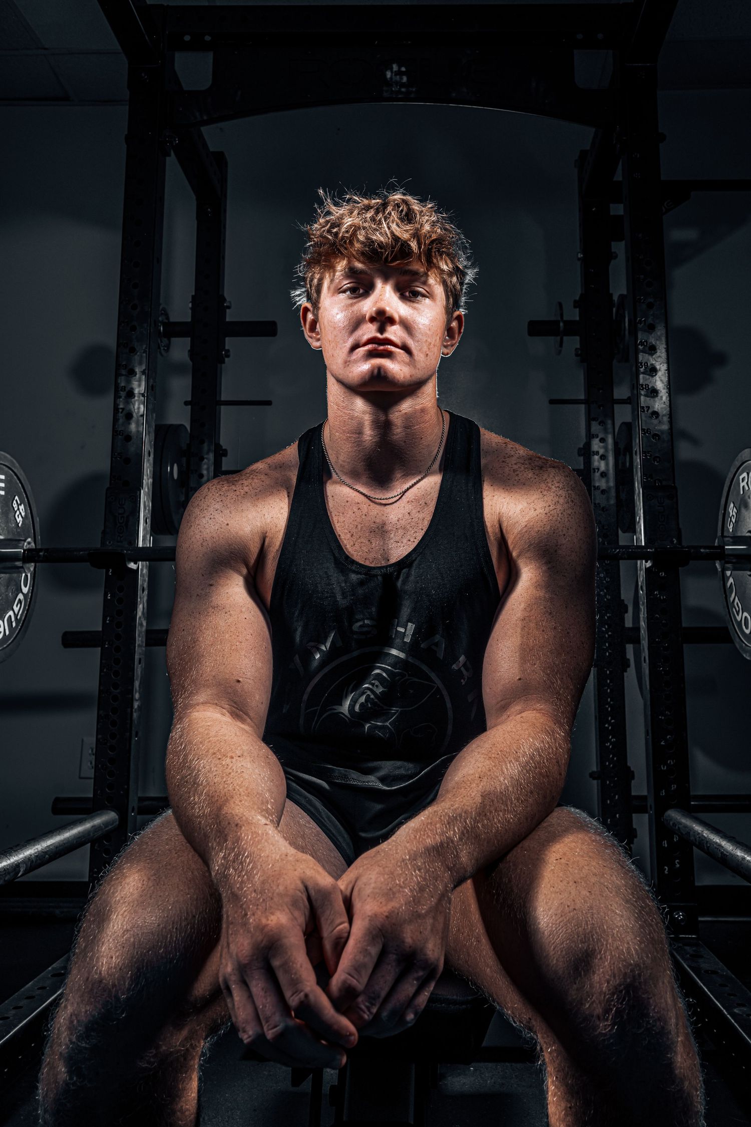 Athletic figure in black tank top sits on weight bench in dramatic gym lighting.