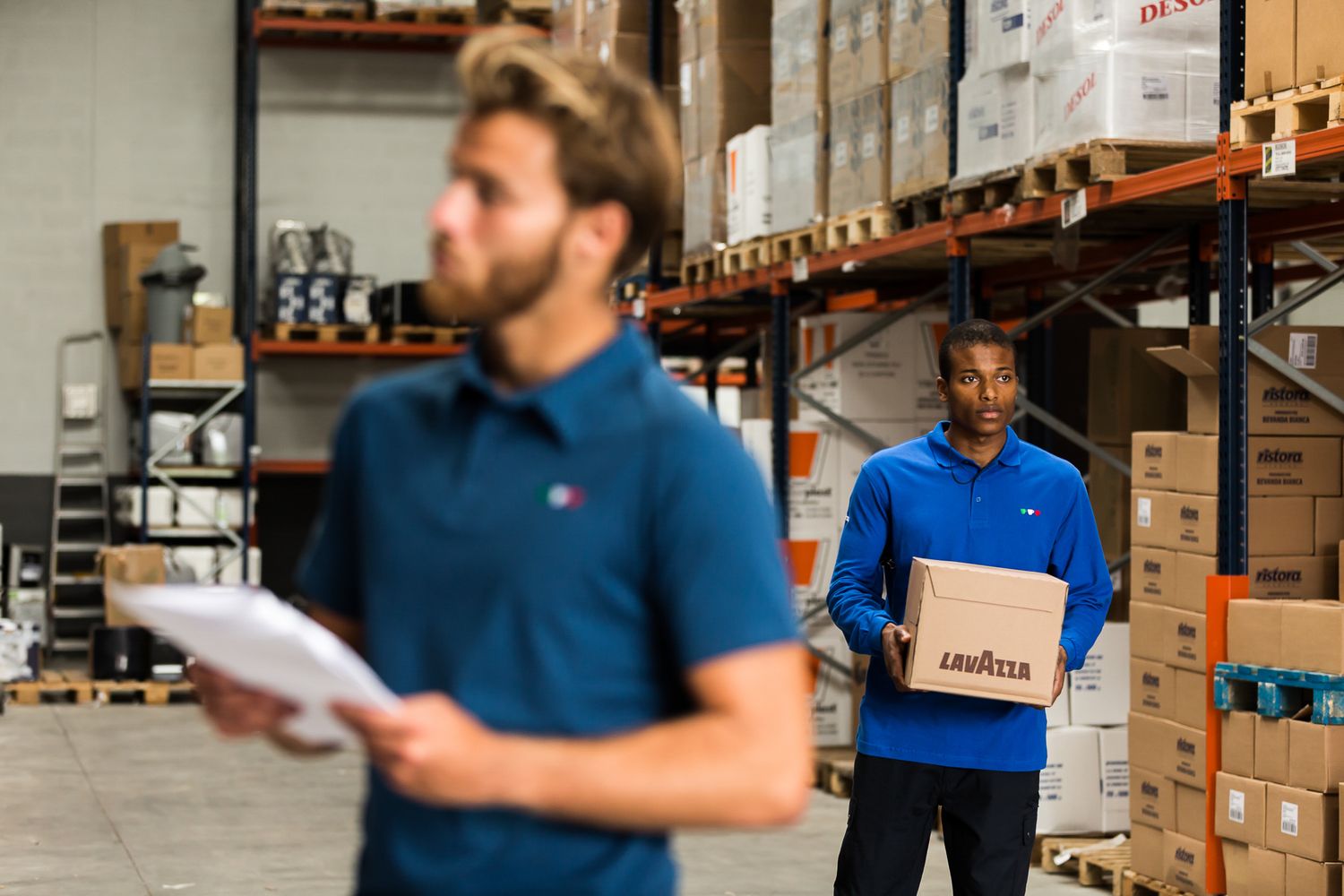 Two workers in blue polo shirts checking inventory in a warehouse storage facility with stacked cardboard boxes on shelves.