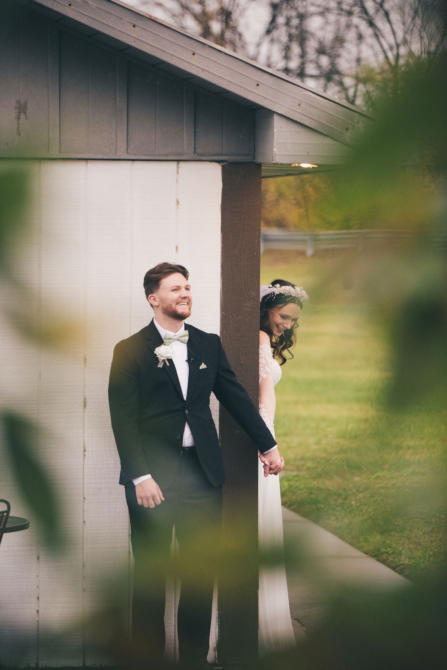 The bride and groom share smiles as the bride peeks at the groom's hand from around the corner of a building.